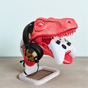 May include: A red dinosaur-shaped headset and controller stand. The dinosaur holds a white controller in its mouth and has black headphones with gold accents and the text "HXS" on its head. The stand is on a rectangular base.