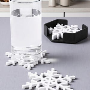 Puede incluir: Un vaso de agua transparente descansa sobre un posavasos blanco en forma de copo de nieve. Se ven posavasos adicionales blancos en forma de copo de nieve, junto con un soporte negro hexagonal para posavasos que contiene m&aacute;s posavasos. Los posavasos son un art&iacute;culo de decoraci&oacute;n del hogar con tem&aacute;tica invernal.