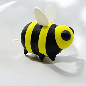 May include: A whimsical bee-shaped toothpick holder. The holder features a yellow and black striped body, black eyes, and small black legs. It has two white, oval-shaped wings and a toothpick extending from the rear.