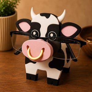 May include: A whimsical cow-shaped eyeglasses holder. The holder is black and white with pink accents for the ears and nose. The cow has a gold-colored ring in its nose and is holding a pair of black-framed glasses. The holder is made of wood.