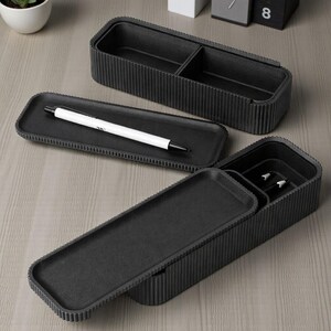 May include: A set of three black desk organizers with a ribbed texture. The set includes a rectangular tray with a white pen, and two rectangular boxes, one with a divided interior. The organizers are made of a matte material.