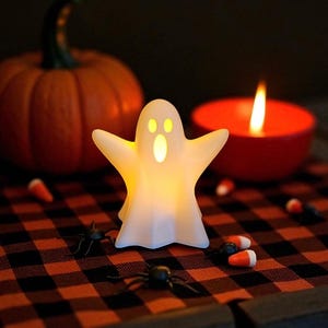 May include: A glowing white ghost-shaped Halloween decoration with outstretched arms. The ghost is lit from within, with a pumpkin and lit candle in the background. Candy corn and plastic spiders are scattered on a red and black checkered surface.