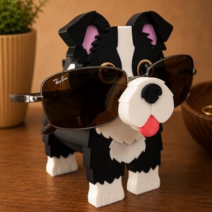 May include: A black, white, and pink dog-shaped eyeglass holder with sunglasses. The dog has a white muzzle, pink ears, and a red tongue. The sunglasses are brown with silver frames and the Ray-Ban logo.