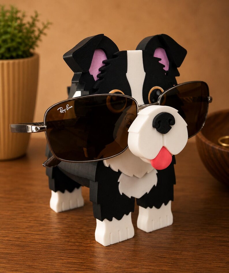 May include: A black, white, and pink dog-shaped eyeglass holder with sunglasses. The dog has a white muzzle, pink ears, and a red tongue. The sunglasses are brown with silver frames and the Ray-Ban logo.