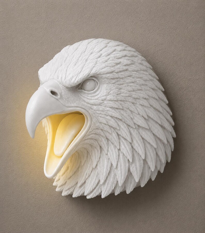 May include: A white eagle head wall sculpture with an open beak, revealing a warm, yellow interior. The sculpture features detailed feather textures and is mounted on a neutral-toned wall. The eagle's eye is visible, and the overall design is a decorative wall accent.