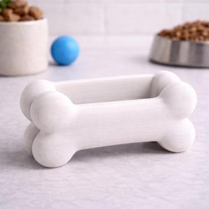 K&ouml;nnte beinhalten: Ein wei&szlig;er, knochenf&ouml;rmiger Halter f&uuml;r Tierleckerlis. Der Halter besteht aus einem festen Material und hat eine glatte, matte Oberfl&auml;che. Ein blauer Ball und eine Sch&uuml;ssel mit Tierfutter befinden sich im Hintergrund.