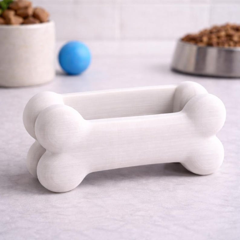 K&ouml;nnte beinhalten: Ein wei&szlig;er, knochenf&ouml;rmiger Halter f&uuml;r Tierleckerlis. Der Halter besteht aus einem festen Material und hat eine glatte, matte Oberfl&auml;che. Ein blauer Ball und eine Sch&uuml;ssel mit Tierfutter befinden sich im Hintergrund.
