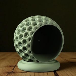 May include: A light green, spherical decorative object with a textured surface and a large opening. The object sits on a matching round base. The surface has a pattern of small, evenly spaced indentations. The background is dark.