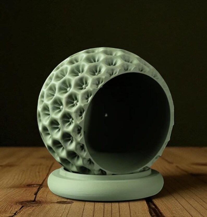 May include: A light green, spherical decorative object with a textured surface and a large opening. The object sits on a matching round base. The surface has a pattern of small, evenly spaced indentations. The background is dark.
