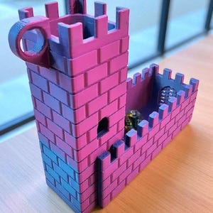 May include: A pink and blue miniature castle with a cylindrical handle. The castle has a rectangular tower and a connecting wall, both with crenellations. A small, dark die is visible inside the wall. The castle is made of plastic.