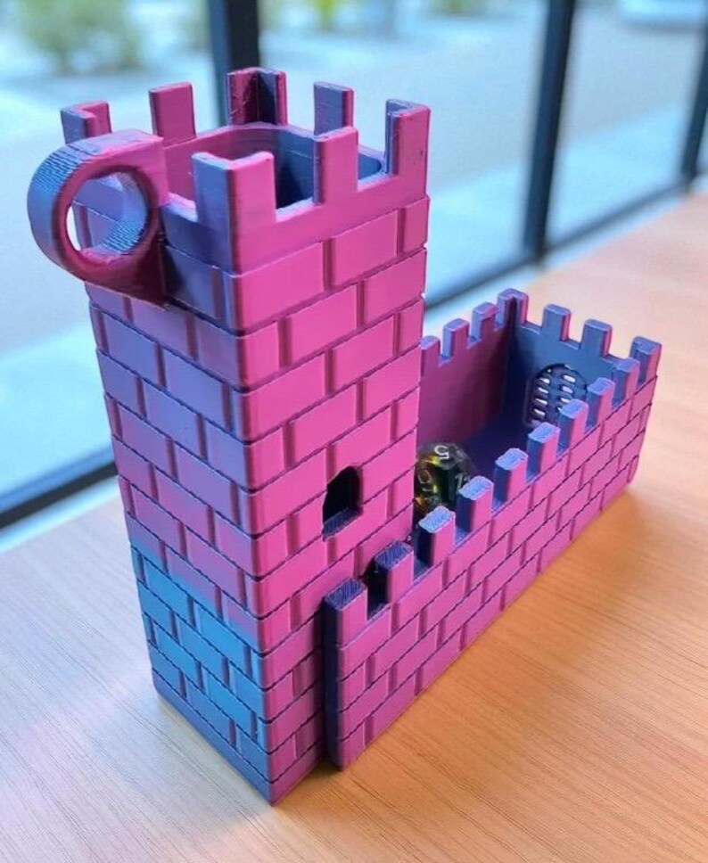 May include: A pink and blue miniature castle with a cylindrical handle. The castle has a rectangular tower and a connecting wall, both with crenellations. A small, dark die is visible inside the wall. The castle is made of plastic.