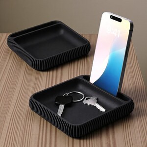 May include: Two black square trays with ribbed sides. One tray holds a smartphone, the other holds keys. The trays are on a wooden surface. The smartphone screen displays the time.