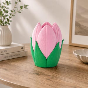 May include: A decorative flower-shaped object in pink and green. The pink petals have a textured finish, and the green base is leaf-like. It sits on a wooden table, with a white vase and books visible in the background.