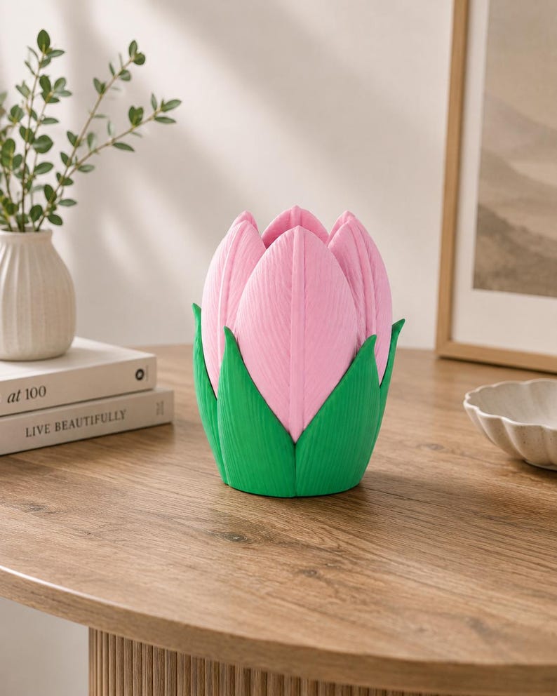 May include: A decorative flower-shaped object in pink and green. The pink petals have a textured finish, and the green base is leaf-like. It sits on a wooden table, with a white vase and books visible in the background.