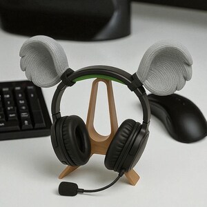 May include: Black headphones with a microphone, resting on a wooden stand. The headphones have gray ear covers shaped like wings. A black computer mouse and keyboard are in the background.