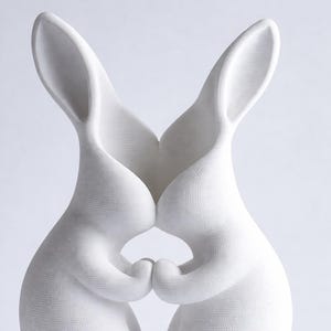 May include: A white ceramic sculpture of two rabbits embracing. The rabbits are depicted with their ears touching and their paws clasped together. The sculpture has a textured surface and a minimalist design.
