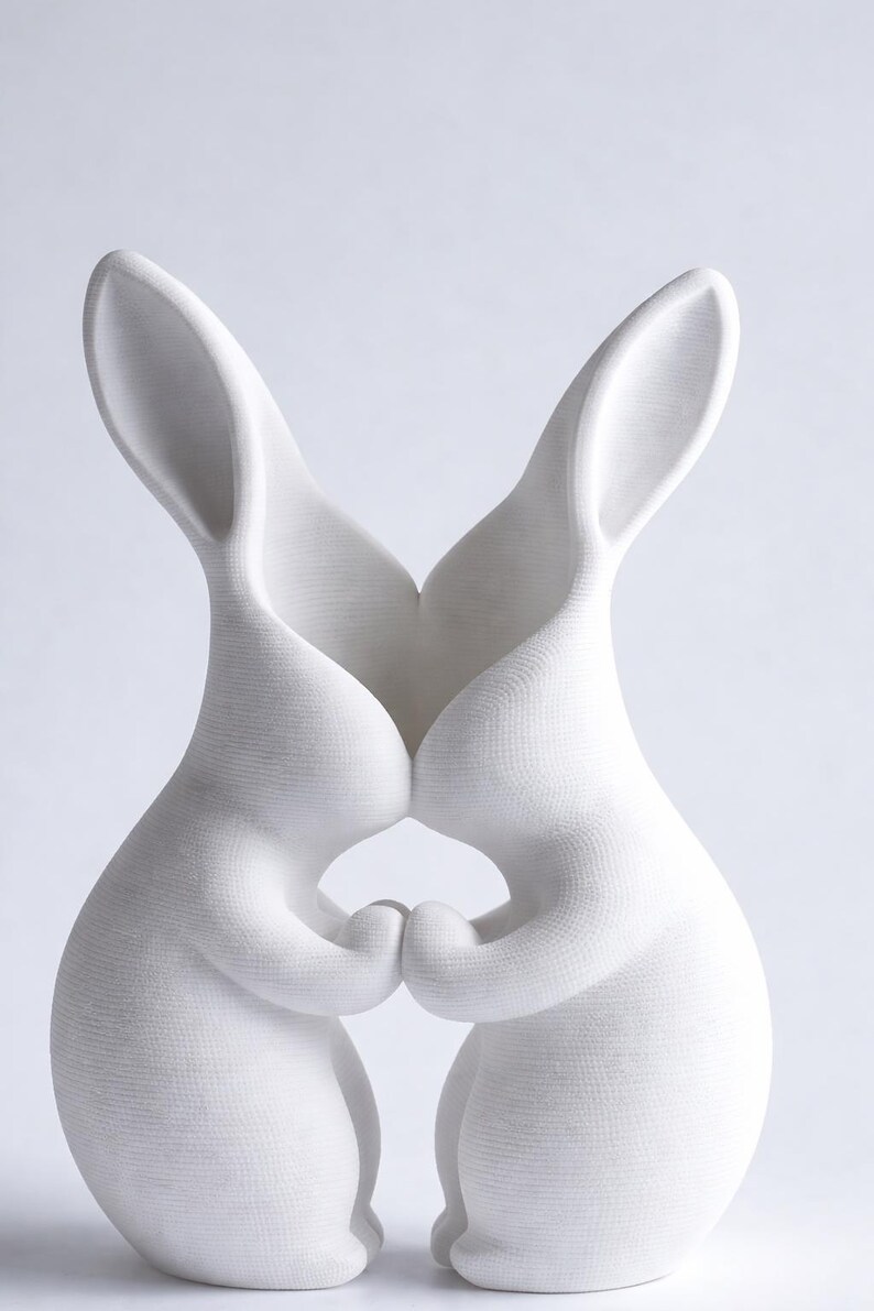 May include: A white ceramic sculpture of two rabbits embracing. The rabbits are depicted with their ears touching and their paws clasped together. The sculpture has a textured surface and a minimalist design.