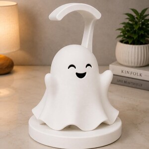 May include: A white ghost-shaped night light with a smiling face and outstretched arms. The lamp has a curved handle and sits on a round base. A small potted plant and a lamp are in the background.