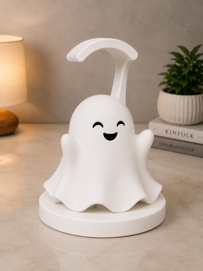 May include: A white ghost-shaped night light with a smiling face and outstretched arms. The lamp has a curved handle and sits on a round base. A small potted plant and a lamp are in the background.