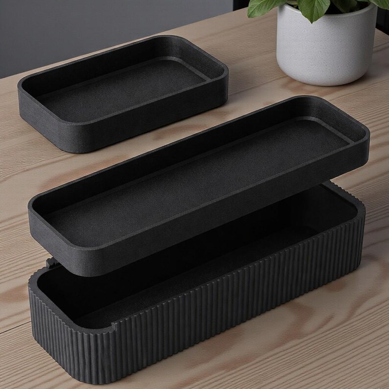May include: Two black rectangular storage trays. One tray is open, revealing a second tray inside. The bottom tray has vertical ribbed detailing. The other tray is a smaller, solid rectangle. The trays are on a light wood surface.