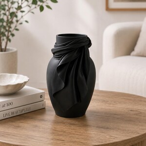 May include: A matte black decorative vase with a draped fabric design around the neck. The vase is cylindrical with a wide opening and sits on a wooden table. A plant and books are in the background.