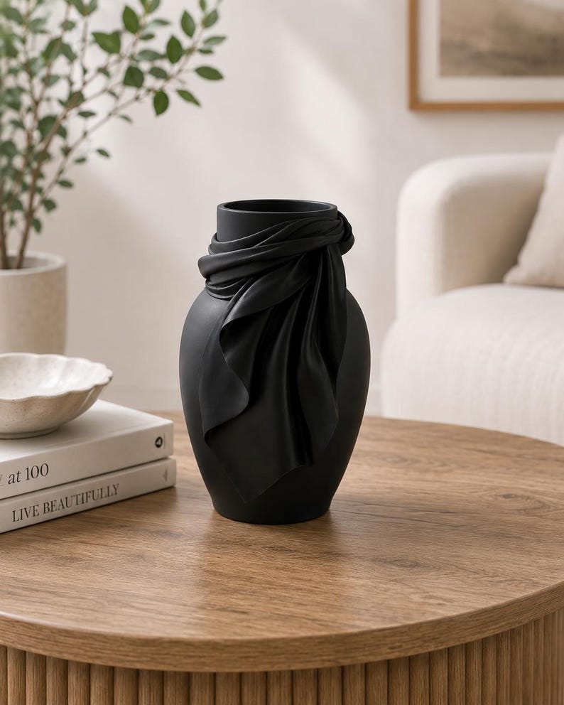May include: A matte black decorative vase with a draped fabric design around the neck. The vase is cylindrical with a wide opening and sits on a wooden table. A plant and books are in the background.