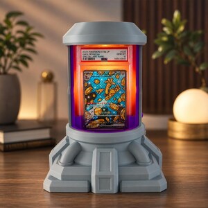 Op de afbeelding: Een grijze, paarse en oranje display met een Pok&eacute;mon-kaart erin. De kaart is een Special Art Rare Gholdengo-kaart. De behuizing heeft een futuristisch ontwerp en wordt verlicht door oranje en paars licht.