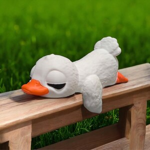 May include: A white ceramic duck figurine with an orange beak and feet, lying on a wooden bench. The duck has closed eyes, suggesting it is sleeping. The background is blurred green grass.