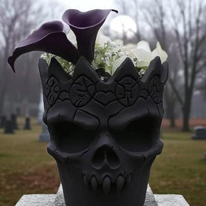 May include: A black skull-shaped plant pot, filled with dark purple calla lilies, white flowers, and greenery. The pot has a crown design around the top. It sits on a stone surface, with a blurred cemetery in the background.