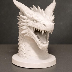 May include: A white dragon head sculpture with open mouth, sharp teeth, and detailed scales. The dragon has prominent horns and a spiked ridge along its back. The sculpture is mounted on a circular base, showcasing a fantasy theme.