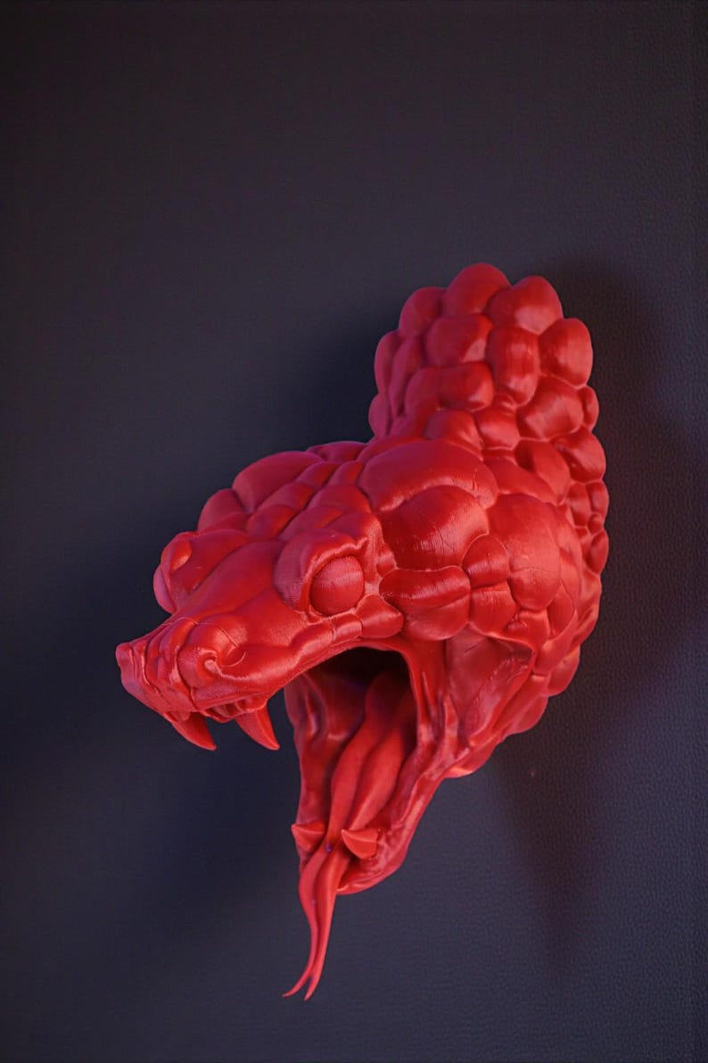 May include: A vibrant red sculpture of a snake's head, with its mouth open, revealing fangs and a forked tongue. The detailed design showcases textured scales and a menacing expression. The sculpture is set against a dark background.