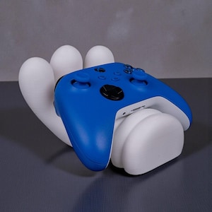 May include: A blue and black video game controller rests on a white, hand-shaped stand. The controller has a sleek design with buttons and joysticks. The stand is a unique, modern accessory for gamers.