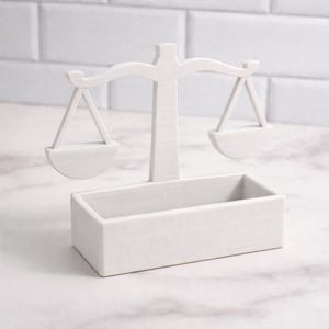 Puede incluir: Una balanza decorativa blanca con base rectangular. La balanza tiene dos bandejas colgantes y un soporte central. El dise&ntilde;o es minimalista y la balanza est&aacute; hecha de un material s&oacute;lido y texturizado. Es un art&iacute;culo decorativo.