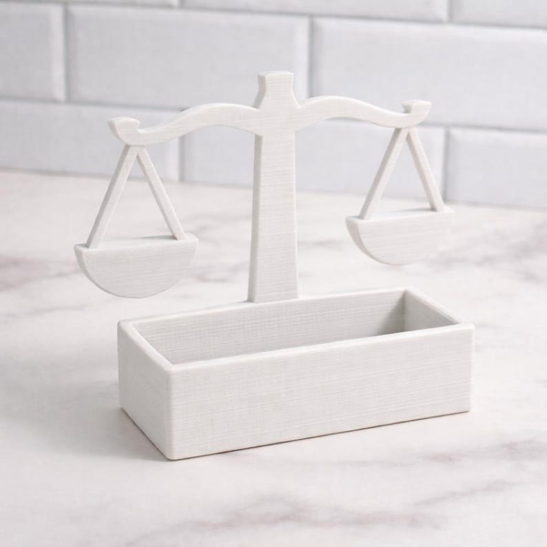 Puede incluir: Una balanza decorativa blanca con base rectangular. La balanza tiene dos bandejas colgantes y un soporte central. El dise&ntilde;o es minimalista y la balanza est&aacute; hecha de un material s&oacute;lido y texturizado. Es un art&iacute;culo decorativo.