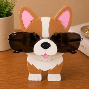 May include: A brown and white corgi-shaped sunglasses holder with pink inner ears and a white snout. The holder is wearing a pair of Ray-Ban sunglasses with a silver frame and dark lenses. The holder is on a wooden surface.
