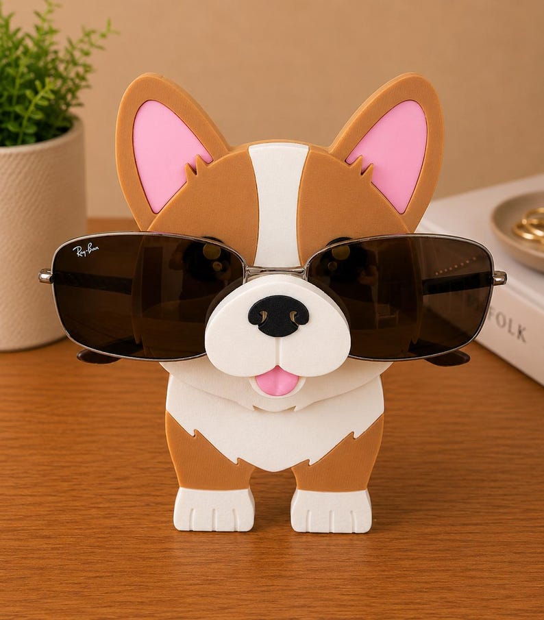 May include: A brown and white corgi-shaped sunglasses holder with pink inner ears and a white snout. The holder is wearing a pair of Ray-Ban sunglasses with a silver frame and dark lenses. The holder is on a wooden surface.