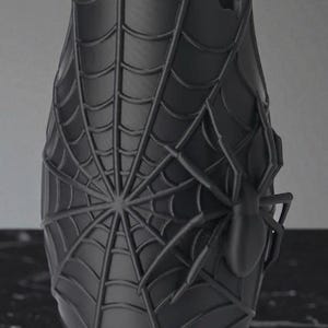 May include: A dark gray vase with a spiderweb design and a spider on the side. The vase has a cylindrical shape with a slightly flared top and a damaged section on the rim. The spiderweb covers most of the vase's surface.