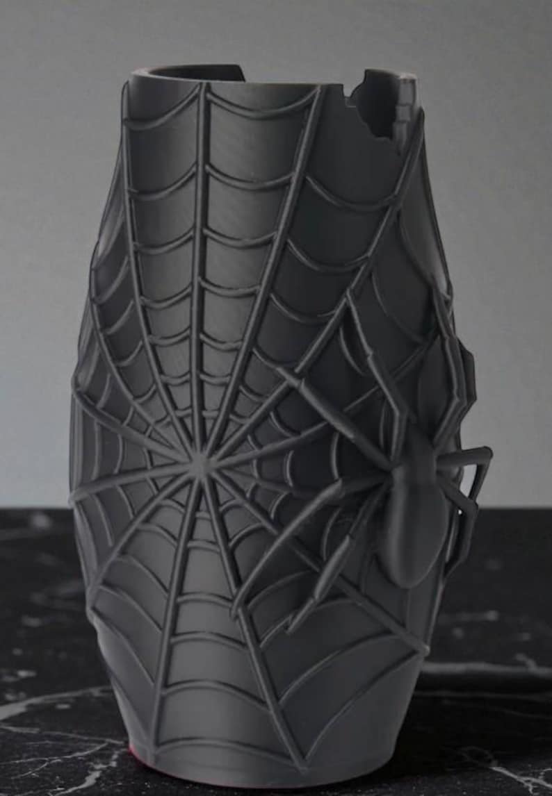 May include: A dark gray vase with a spiderweb design and a spider on the side. The vase has a cylindrical shape with a slightly flared top and a damaged section on the rim. The spiderweb covers most of the vase's surface.