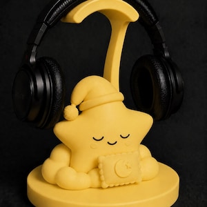 May include: A yellow star-shaped headphone stand with a sleeping cap and a pillow with a crescent moon design. Black headphones rest on the stand. The stand is on a round base, all against a black background.