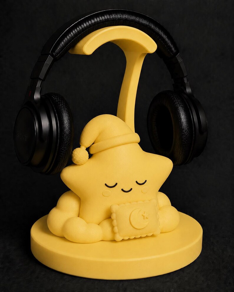 May include: A yellow star-shaped headphone stand with a sleeping cap and a pillow with a crescent moon design. Black headphones rest on the stand. The stand is on a round base, all against a black background.