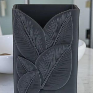 May include: A dark gray, rectangular container with a leaf design. The leaves are embossed on the front, creating a textured surface. The container is likely for holding items in a bathroom or on a vanity.