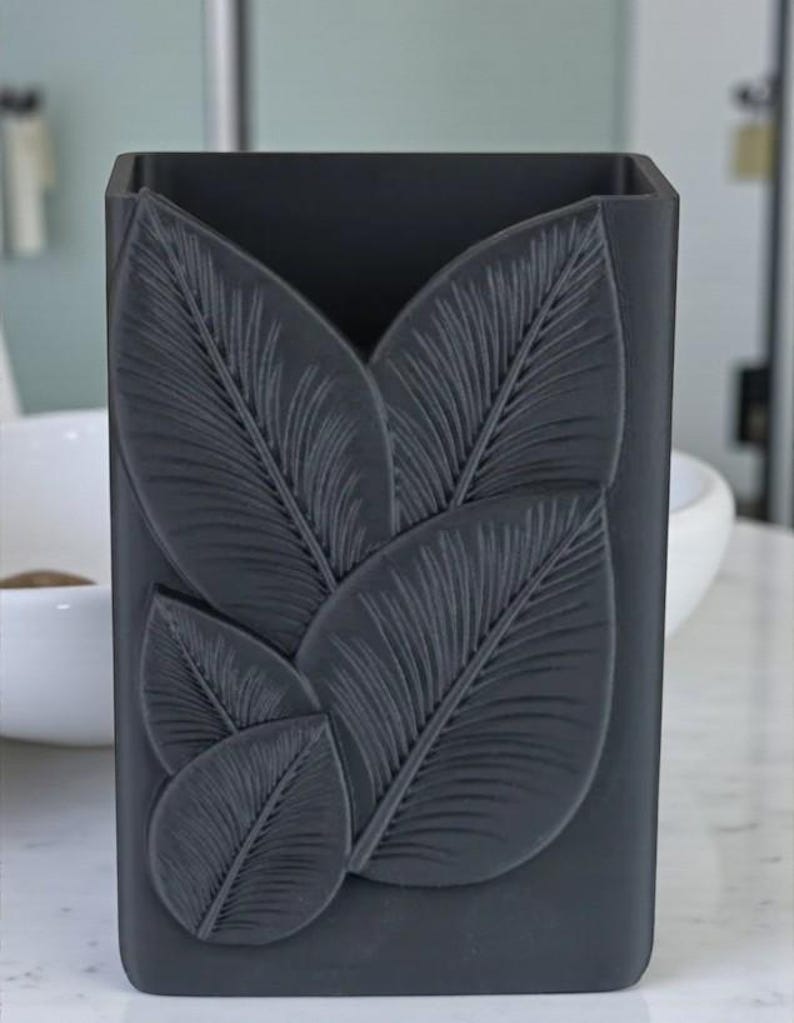 May include: A dark gray, rectangular container with a leaf design. The leaves are embossed on the front, creating a textured surface. The container is likely for holding items in a bathroom or on a vanity.