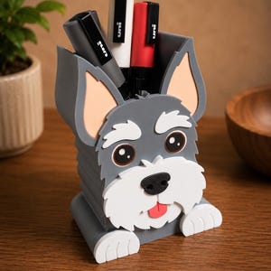 May include: A gray dog-shaped desk organizer with a white muzzle, ears, and paws. The organizer holds several black, white, and red pens. The dog has a pink tongue sticking out and brown eyes. The organizer is on a wooden surface.