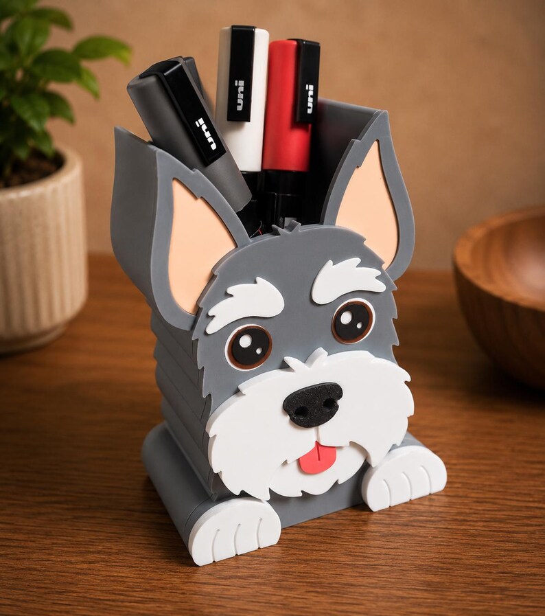 May include: A gray dog-shaped desk organizer with a white muzzle, ears, and paws. The organizer holds several black, white, and red pens. The dog has a pink tongue sticking out and brown eyes. The organizer is on a wooden surface.