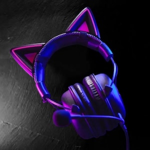 May include: Purple and pink cat ear headphones with a microphone. The headphones have a black headband and earcups.