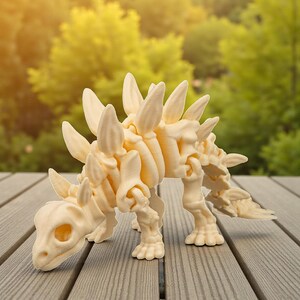 May include: A cream-colored stegosaurus dinosaur figurine with prominent dorsal plates and a detailed skeletal structure. The dinosaur is posed on a wooden surface, with a blurred green background of trees.