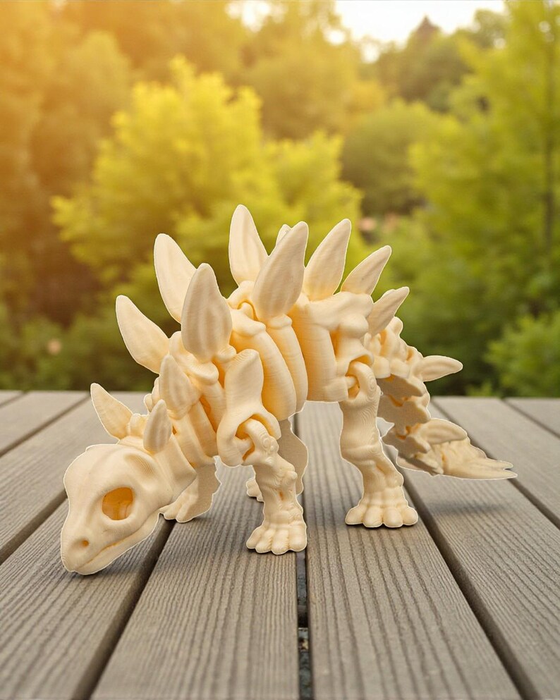 May include: A cream-colored stegosaurus dinosaur figurine with prominent dorsal plates and a detailed skeletal structure. The dinosaur is posed on a wooden surface, with a blurred green background of trees.
