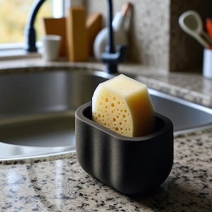 May include: A black sponge holder with a light yellow sponge inside. The holder has a curved design and is sitting on a speckled countertop near a kitchen sink. The sponge has many small holes.