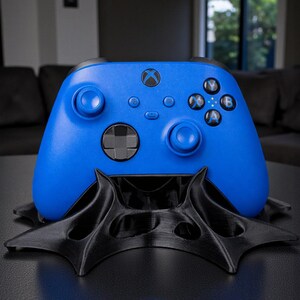 May include: A vibrant blue Xbox controller rests on a black, stylized stand. The controller features the Xbox logo and various buttons. The stand has a unique, geometric design, creating a modern aesthetic.