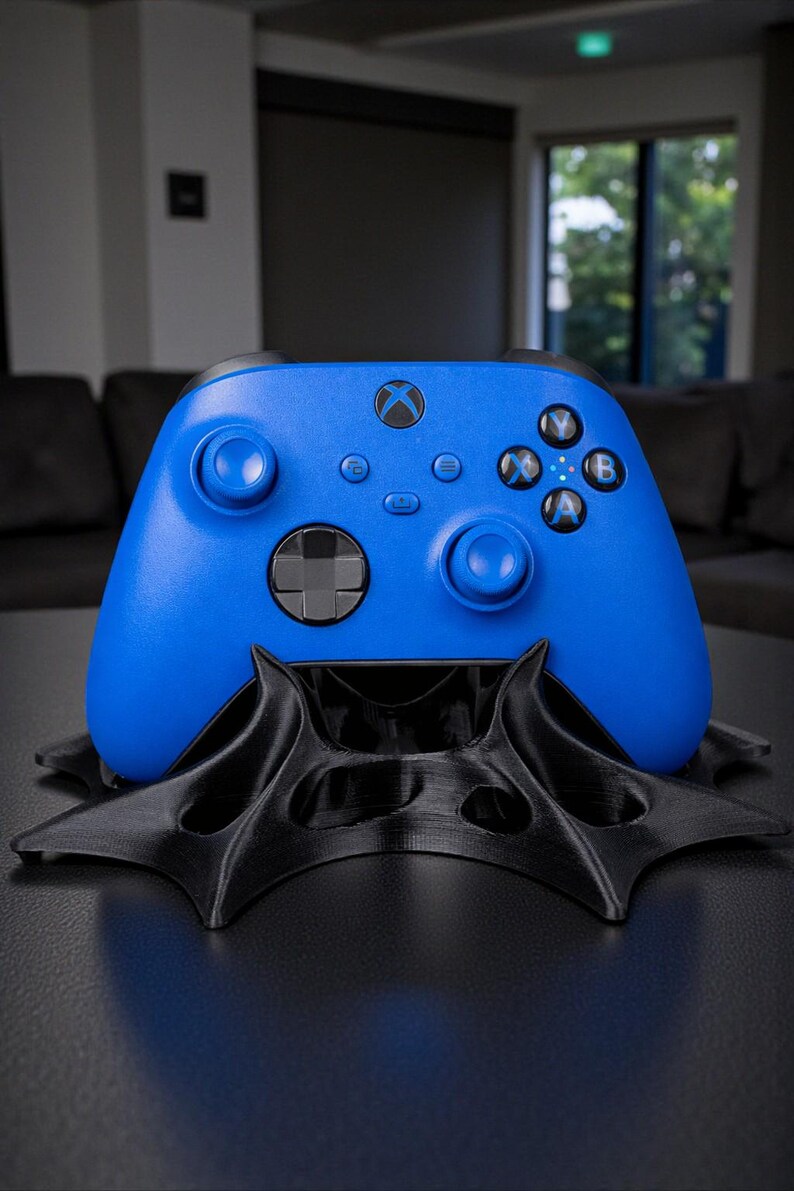 May include: A vibrant blue Xbox controller rests on a black, stylized stand. The controller features the Xbox logo and various buttons. The stand has a unique, geometric design, creating a modern aesthetic.